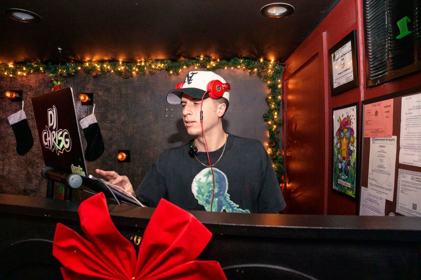 DJ Chris G spinning at a festive bar DJ booth with holiday lights